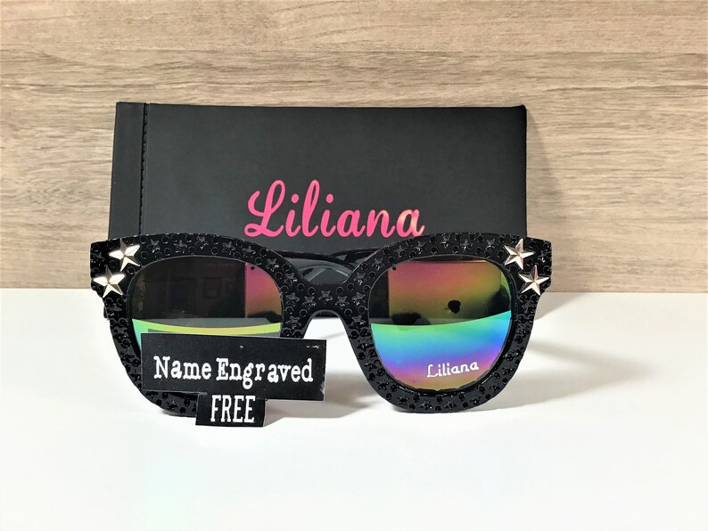 Puede incluir: Gafas de sol negras con lentes iridiscentes y detalles de estrellas plateadas. Las gafas tienen el nombre "Liliana" grabado en la lente derecha y una etiqueta que dice "Nombre grabado GRATIS".