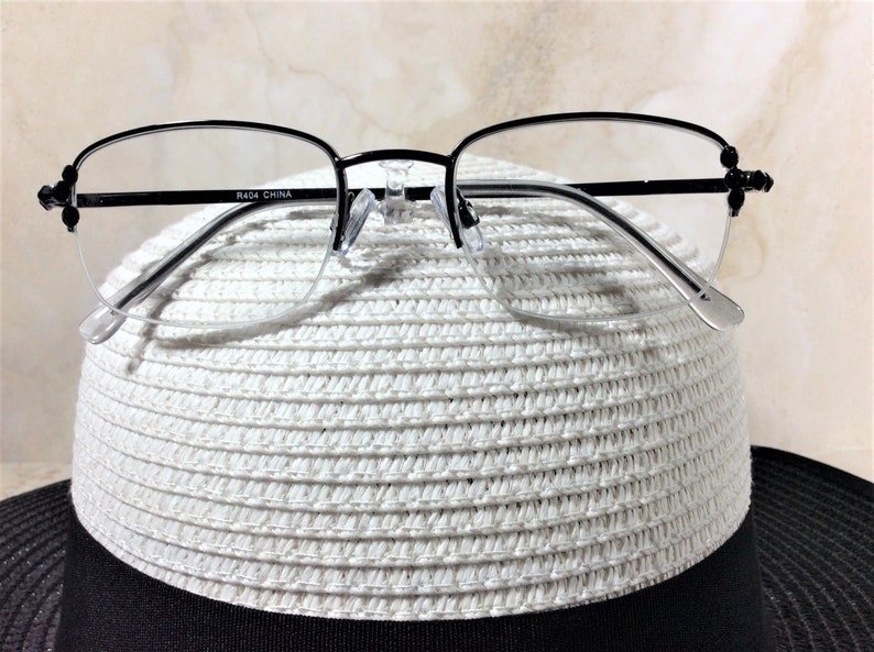 May include: Black half-rim eyeglasses with clear lenses rest on a white woven hat with a black brim. The glasses have a delicate design with small decorative elements on the temples. The text "R404 CHINA" is printed on the frame.