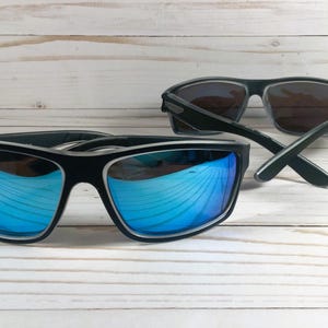 K&ouml;nnte beinhalten: Zwei Sonnenbrillen, eine mit blauen verspiegelten Gl&auml;sern und eine mit dunklen Gl&auml;sern. Beide Brillen haben schwarze Rahmen.