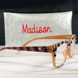 Op de afbeelding: Oranje montuurbril met decoratieve roze en zwarte edelstenen. De veren hebben een bruin en tan patroon. Achter de bril staat een zilveren etui met de naam "Madison" in rode letters.