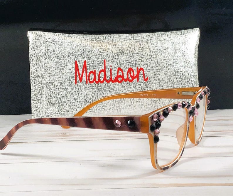 Op de afbeelding: Oranje montuurbril met decoratieve roze en zwarte edelstenen. De veren hebben een bruin en tan patroon. Achter de bril staat een zilveren etui met de naam "Madison" in rode letters.