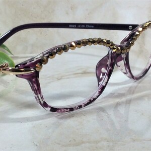 May include: Purple and clear patterned reading glasses with a gold-tone metallic accent at the temple hinge. The upper rims are embellished with a row of faceted bronze-coloured beads. The inner temple arm is printed with R625 +2.00 China.