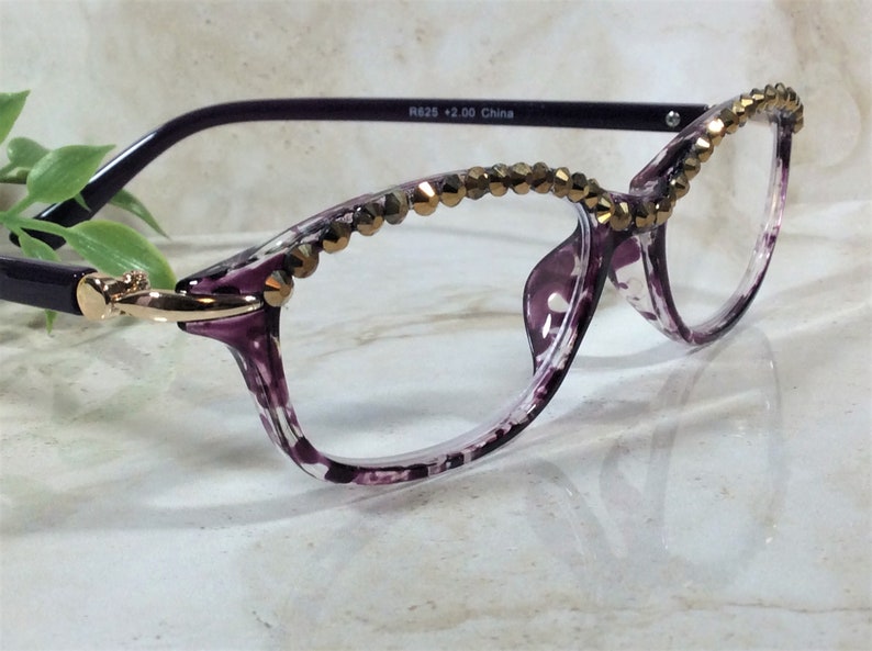 May include: Purple and clear patterned reading glasses with a gold-tone metallic accent at the temple hinge. The upper rims are embellished with a row of faceted bronze-coloured beads. The inner temple arm is printed with R625 +2.00 China.