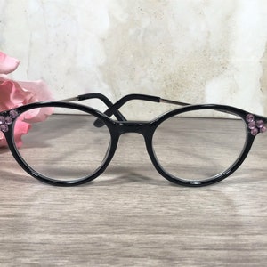 Puede incluir: Gafas negras con lentes transparentes. Las monturas est&aacute;n adornadas con peque&ntilde;os brillantes rosas en las sienes. Las gafas se colocan sobre un fondo neutro con una flor rosa en la esquina.