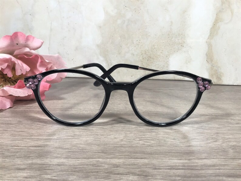 Puede incluir: Gafas negras con lentes transparentes. Las monturas est&aacute;n adornadas con peque&ntilde;os brillantes rosas en las sienes. Las gafas se colocan sobre un fondo neutro con una flor rosa en la esquina.