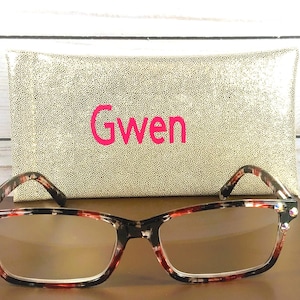 Pu&ograve; includere: Occhiali rettangolari con motivo tartarugato e lenti trasparenti. Sono presentati con un astuccio dorato con paillettes con il nome "Gwen" in rosa. Lo sfondo &egrave; una superficie di legno chiaro.