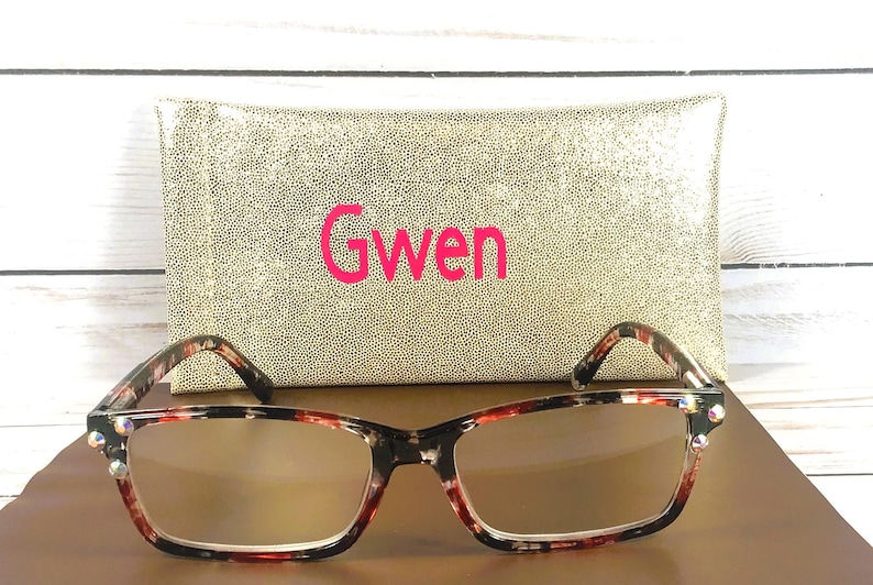 Pu&ograve; includere: Occhiali rettangolari con motivo tartarugato e lenti trasparenti. Sono presentati con un astuccio dorato con paillettes con il nome "Gwen" in rosa. Lo sfondo &egrave; una superficie di legno chiaro.
