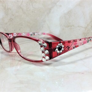 Peut inclure: Lunettes &agrave; monture rouge avec verres transparents. Les montures sont orn&eacute;es de strass transparents et d'un motif floral. Une gemme noire est sertie dans le coin de la monture. Les branches des lunettes ont un motif floral rose et blanc.