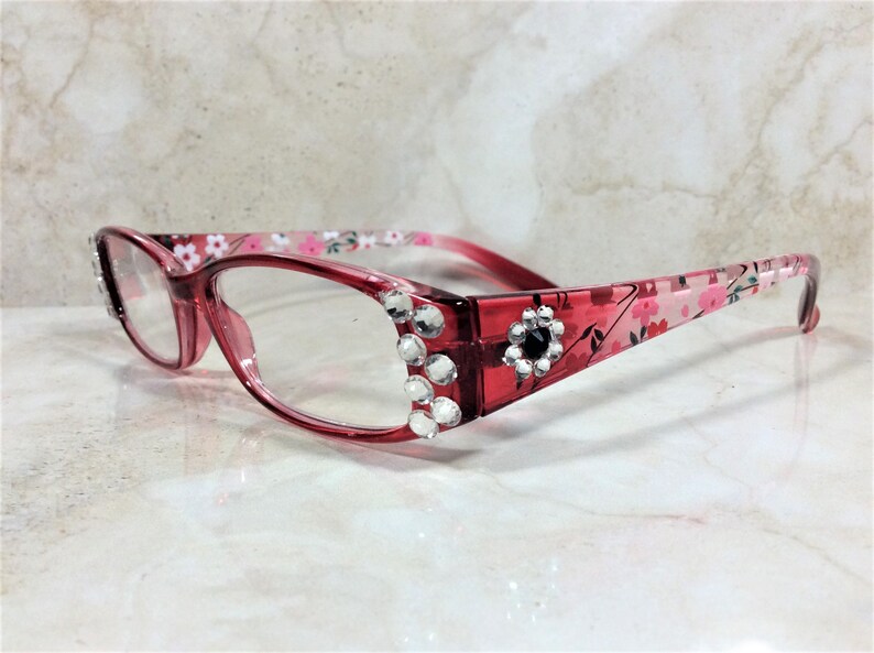 Peut inclure: Lunettes &agrave; monture rouge avec verres transparents. Les montures sont orn&eacute;es de strass transparents et d'un motif floral. Une gemme noire est sertie dans le coin de la monture. Les branches des lunettes ont un motif floral rose et blanc.