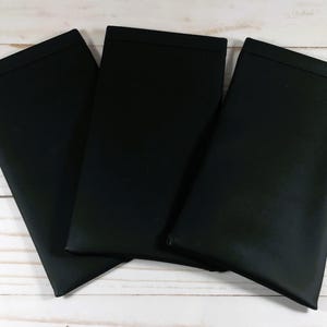 May include: Three black fabric placemats, folded and stacked on a white wooden surface.
