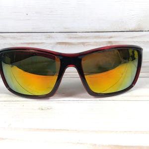 May include: Sport sunglasses with a black frame and red accents. The lenses have a mirrored finish with yellow and orange hues. The sunglasses are set against a white wooden background.