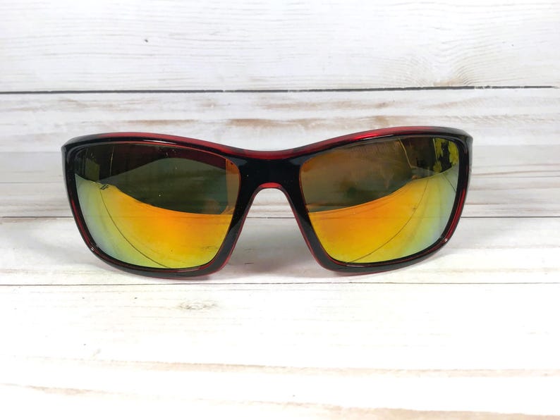 May include: Sport sunglasses with a black frame and red accents. The lenses have a mirrored finish with yellow and orange hues. The sunglasses are set against a white wooden background.
