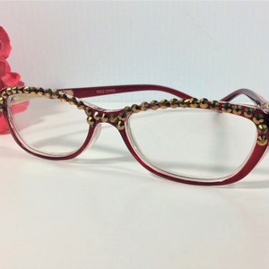 May include: Red-framed spectacles with clear lenses, decorated with gold-coloured embellishments along the top rim. The spectacles have a cat-eye shape and the text "8622 CHINA" is printed on the temple.