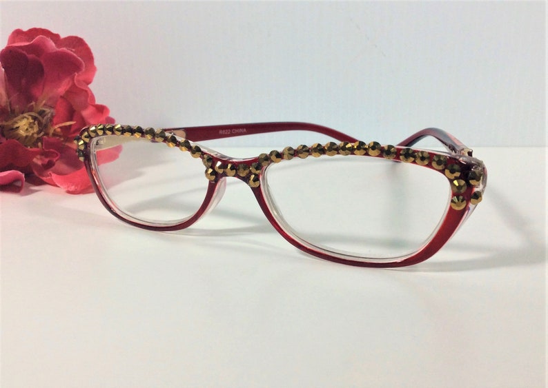 May include: Red-framed spectacles with clear lenses, decorated with gold-coloured embellishments along the top rim. The spectacles have a cat-eye shape and the text "8622 CHINA" is printed on the temple.