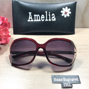 Op de afbeelding: Bordeauxkleurige zonnebril met een gouden rand en een zwarte etui. De etui heeft de naam "Amelia" en een witte madeliefje erop gedrukt. De zonnebril heeft een "Protection" label aan de zijkant.