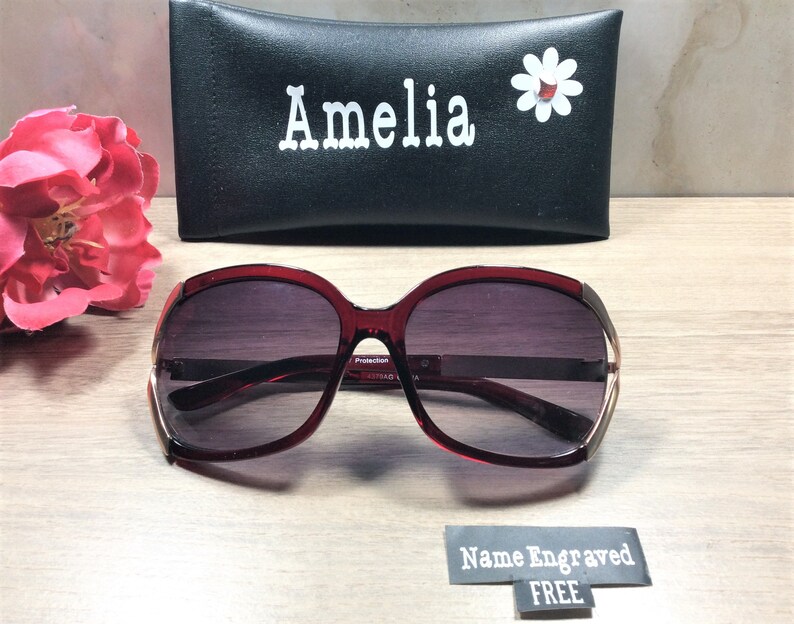 Op de afbeelding: Bordeauxkleurige zonnebril met een gouden rand en een zwarte etui. De etui heeft de naam "Amelia" en een witte madeliefje erop gedrukt. De zonnebril heeft een "Protection" label aan de zijkant.