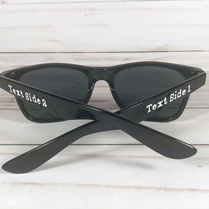K&ouml;nnte beinhalten: Schwarze Sonnenbrille mit dunklen Gl&auml;sern. Die B&uuml;gel der Sonnenbrille sind gekreuzt, mit dem Text "Text Side 1" und "Text Side 2" in Wei&szlig; auf den B&uuml;geln. Die Sonnenbrille steht vor einem hellen Holzgrund.