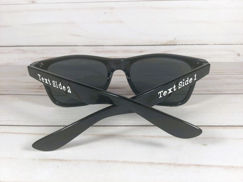 K&ouml;nnte beinhalten: Schwarze Sonnenbrille mit dunklen Gl&auml;sern. Die B&uuml;gel der Sonnenbrille sind gekreuzt, mit dem Text "Text Side 1" und "Text Side 2" in Wei&szlig; auf den B&uuml;geln. Die Sonnenbrille steht vor einem hellen Holzgrund.