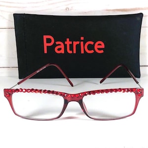 May include: Red-framed eyeglasses with a decorative crystal border, resting in front of a black case. The case has the name "Patrice" in red lettering. The background is a white wooden surface.