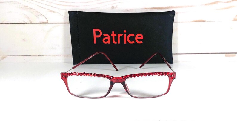 May include: Red-framed eyeglasses with a decorative crystal border, resting in front of a black case. The case has the name "Patrice" in red lettering. The background is a white wooden surface.