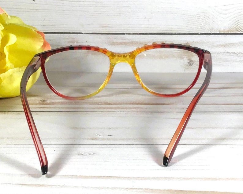 Puede incluir: Gafas con una montura degradada que pasa de un rojo intenso en los bordes exteriores a un amarillo brillante en el puente. Las monturas tienen un acabado transl&uacute;cido e incluyen peque&ntilde;os tachones decorativos a lo largo del borde superior.
