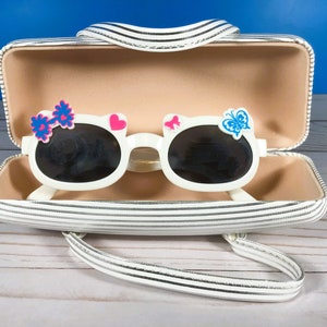 May include: White sunglasses with black lenses, decorated with a pink heart, blue butterfly, and a blue and pink flower. The sunglasses are inside a striped case with a handle. The case is white with black stripes.
