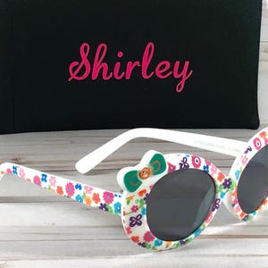 May include: White sunglasses with a floral pattern and a green bow on the side. The sunglasses have a black case with the name "Shirley" printed on it.