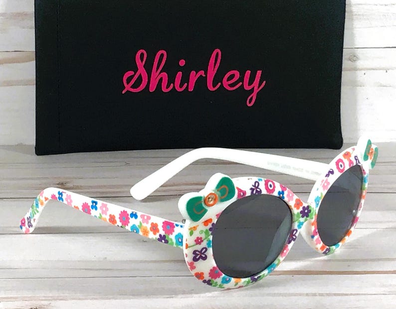 May include: White sunglasses with a floral pattern and a green bow on the side. The sunglasses have a black case with the name "Shirley" printed on it.