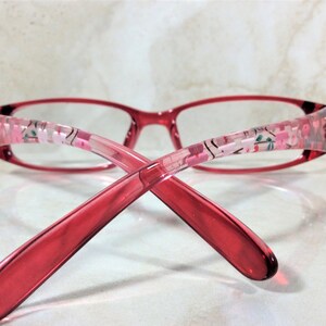 Peut inclure: Lunettes rouges avec des motifs floraux sur les branches. Les montures sont rouge translucide, et les branches pr&eacute;sentent un motif floral rose et blanc. Les lunettes sont de forme rectangulaire, sur fond clair.