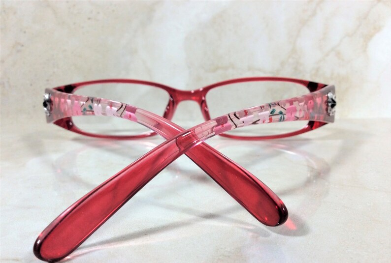 Peut inclure: Lunettes rouges avec des motifs floraux sur les branches. Les montures sont rouge translucide, et les branches pr&eacute;sentent un motif floral rose et blanc. Les lunettes sont de forme rectangulaire, sur fond clair.