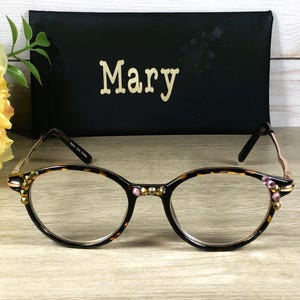 May include: Oval-shaped eyeglasses with a tortoiseshell pattern frame, adorned with pink and gold embellishments. A black case with the name "Mary" in gold lettering is behind the glasses. A yellow flower and green leaves are in the background.