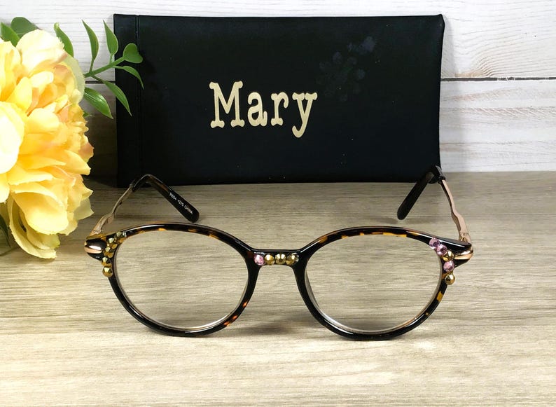 May include: Oval-shaped eyeglasses with a tortoiseshell pattern frame, adorned with pink and gold embellishments. A black case with the name "Mary" in gold lettering is behind the glasses. A yellow flower and green leaves are in the background.