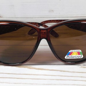 May include: Brown sunglasses with a cat-eye shape. The frames are a dark brown color with small rhinestones along the top edge. The lenses are a brown tint, and a "Polarized" sticker is on the right lens.