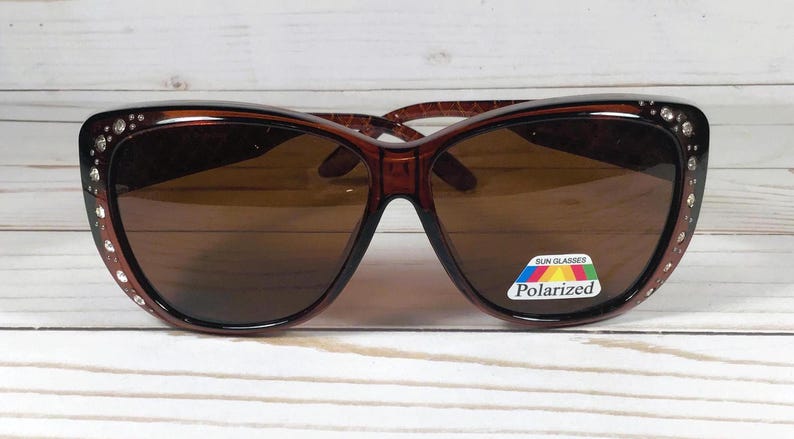 May include: Brown sunglasses with a cat-eye shape. The frames are a dark brown color with small rhinestones along the top edge. The lenses are a brown tint, and a "Polarized" sticker is on the right lens.