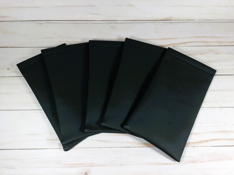 May include: Six black leatherette eyeglass cases arranged in a fan shape on a white wooden surface.