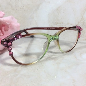 May include: A pair of eyeglasses with a gradient frame, transitioning from brown to green. The frames are adorned with pink rhinestones at the temples and green rhinestones at the bridge. The text "R291 +2.25 China" is printed on the frame.