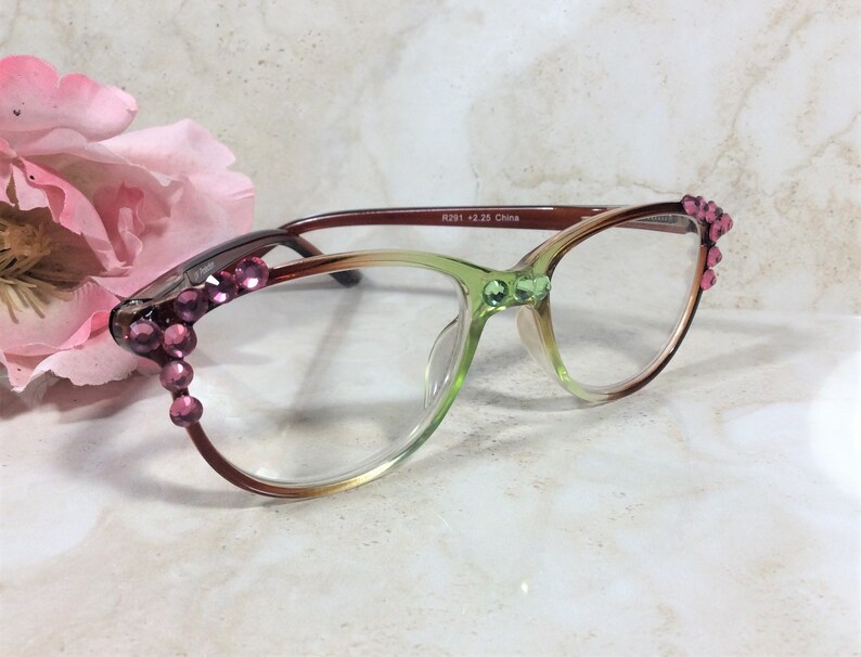 May include: A pair of eyeglasses with a gradient frame, transitioning from brown to green. The frames are adorned with pink rhinestones at the temples and green rhinestones at the bridge. The text "R291 +2.25 China" is printed on the frame.