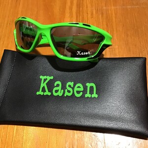 May include: Green and black sunglasses with a black case. The case has the name "Kasen" printed on it in green.