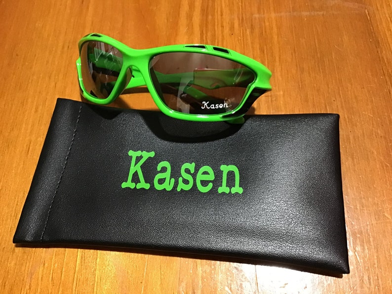 May include: Green and black sunglasses with a black case. The case has the name "Kasen" printed on it in green.