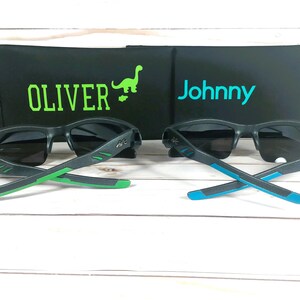 K&ouml;nnte beinhalten: Zwei Paar schwarze Sonnenbrillen mit gr&uuml;nen und blauen Akzenten an den B&uuml;geln. Die Sonnenbrillen liegen auf einer Holzoberfl&auml;che. Die Sonnenbrillen liegen vor zwei schwarzen Etuis mit den Namen "Oliver" und "Johnny" darauf.