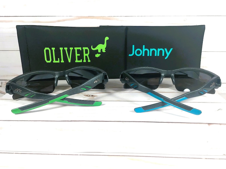 K&ouml;nnte beinhalten: Zwei Paar schwarze Sonnenbrillen mit gr&uuml;nen und blauen Akzenten an den B&uuml;geln. Die Sonnenbrillen liegen auf einer Holzoberfl&auml;che. Die Sonnenbrillen liegen vor zwei schwarzen Etuis mit den Namen "Oliver" und "Johnny" darauf.