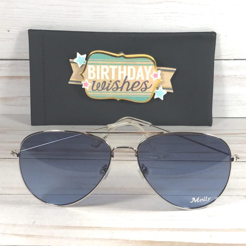 May include: Silver aviator sunglasses with gradient blue lenses, in front of a black case. The case has a decorative label with the words "Birthday Wishes". The name "Molly" is printed on the right lens.