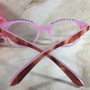 K&ouml;nnte beinhalten: Rosa Cat-Eye-Brille mit dunkler Zierleiste am oberen Rand. Die B&uuml;gel haben ein braunes und rosa Marmormuster. Die Brille liegt vor einem Marmorhintergrund mit einer rosa Blume.