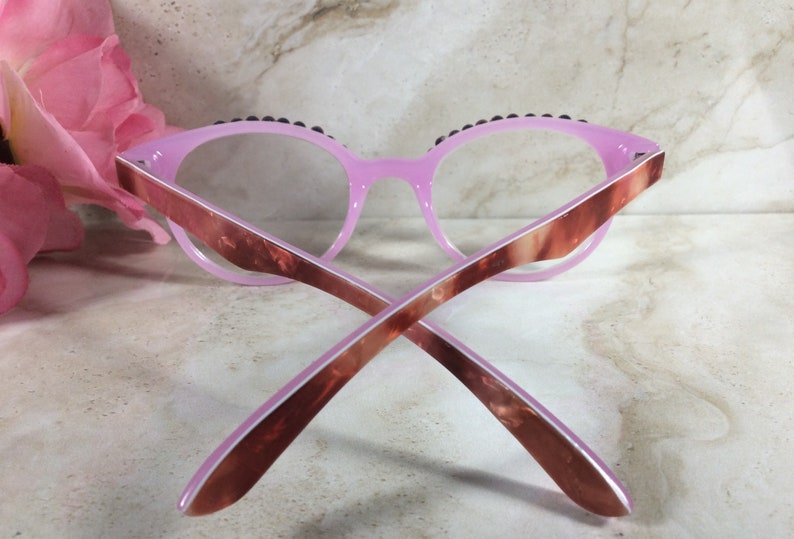 K&ouml;nnte beinhalten: Rosa Cat-Eye-Brille mit dunkler Zierleiste am oberen Rand. Die B&uuml;gel haben ein braunes und rosa Marmormuster. Die Brille liegt vor einem Marmorhintergrund mit einer rosa Blume.