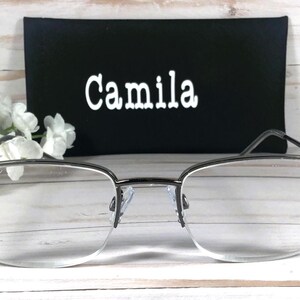 May include: Silver-framed eyeglasses with clear lenses and decorative purple accents on the temples. A black case with the name "Camila" in white lettering is behind the glasses. White flowers are on the left side.