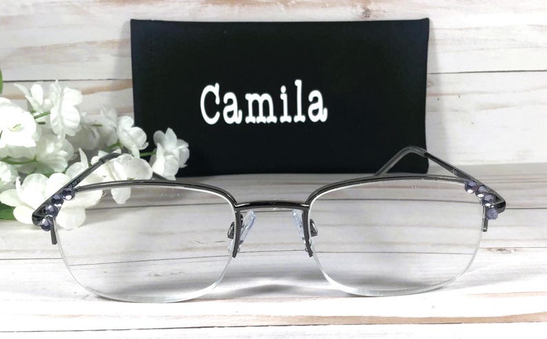 May include: Silver-framed eyeglasses with clear lenses and decorative purple accents on the temples. A black case with the name "Camila" in white lettering is behind the glasses. White flowers are on the left side.