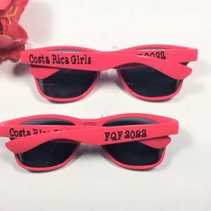 May include: Two pairs of pink plastic sunglasses with black lenses. The top pair has the text "Costa Rica Girls" on the arm. The bottom pair has the text "FQf 2022" on the arm.