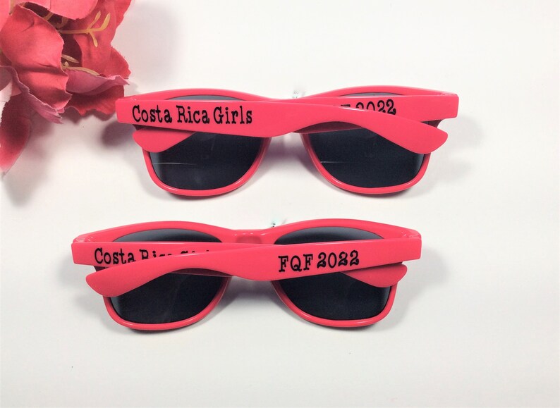 May include: Two pairs of pink plastic sunglasses with black lenses. The top pair has the text "Costa Rica Girls" on the arm. The bottom pair has the text "FQf 2022" on the arm.