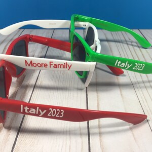 Puede incluir: Tres pares de gafas de sol con monturas blancas y lentes de colores. Las gafas de sol est&aacute;n apiladas unas encima de otras. El par superior es verde, el par del medio es rojo y el par inferior es rojo. Las gafas de sol tienen texto blanco en las patillas que dice "Italia 2023" y "Moore Family".