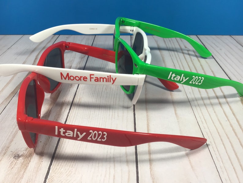 Puede incluir: Tres pares de gafas de sol con monturas blancas y lentes de colores. Las gafas de sol est&aacute;n apiladas unas encima de otras. El par superior es verde, el par del medio es rojo y el par inferior es rojo. Las gafas de sol tienen texto blanco en las patillas que dice "Italia 2023" y "Moore Family".